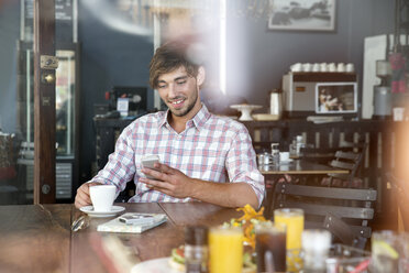 Lächelnder junger Mann in einem Café, der auf sein Handy schaut - TOYF000518