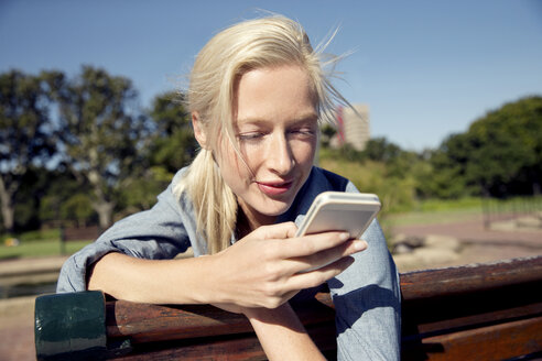 Junge Frau auf Parkbank schaut auf Handy - TOYF000558