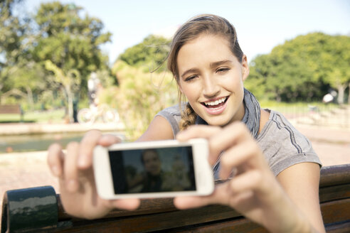 Glückliche junge Frau auf einer Parkbank, die ein Selfie macht - TOYF000559