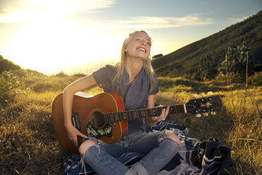 Glückliche junge Frau spielt Gitarre auf einer Decke auf einer Wiese - TOYF000599