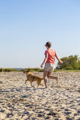 Germany, Kiel, woman running with her dog on sandy beach - JFEF000673