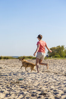 Germany, Kiel, woman running with her dog on sandy beach - JFEF000673