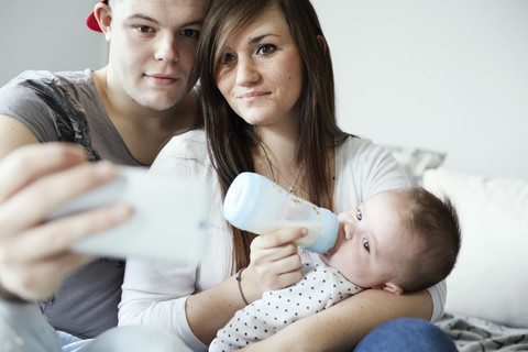 Junger Vater macht Selfie mit Mutter, die ihr Baby zu Hause mit der Flasche füttert, lizenzfreies Stockfoto