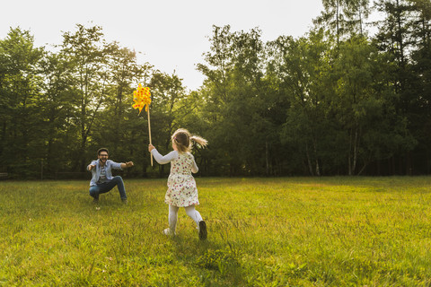 Mädchen mit Papierwindmühle läuft auf der Wiese auf den Vater zu, lizenzfreies Stockfoto