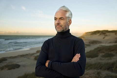 South Africa, portrait of white haired man wearing turtleneck standing on beach dunes before sunrise stock photo