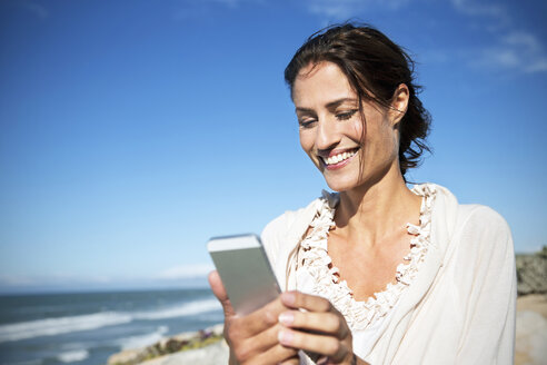 Südafrika, Porträt einer lächelnden Frau, die auf ihr Smartphone schaut, vor dem Meer - TOYF000766