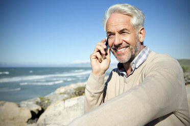 South Africa, portrait of smiling man sitting on rocks telephoning with smartphone - TOYF000807