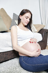 Pregnant woman sitting on the floor of her living room holding her belly - SEGF000369