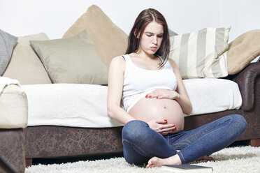 Schwangere Frau sitzt auf dem Boden ihres Wohnzimmers und hält ihren Bauch - SEGF000370