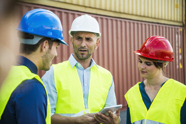 Woman and two men with safety helmets talking at container port - UUF004466