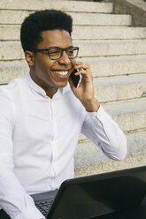 Lächelnder junger Mann sitzt auf einer Treppe und benutzt Handy und Laptop - ABZF000066