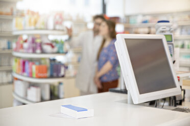 Rezept und Medikament liegen auf dem Tresen einer Apotheke, während der Apotheker im Hintergrund eine Kundin berät - FKF001100