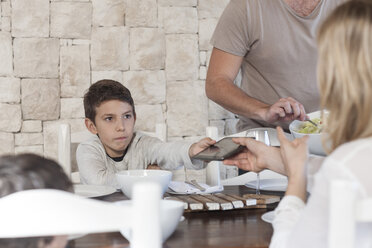 Familie beim Mittagessen am Esstisch mit einem Jungen, der seiner Mutter ein digitales Tablet überreicht - ZEF005377