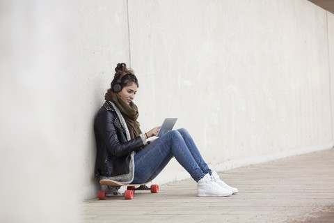 Junge Frau sitzt auf ihrem Longboard mit digitalem Tablet und hört Musik mit Kopfhörern, lizenzfreies Stockfoto