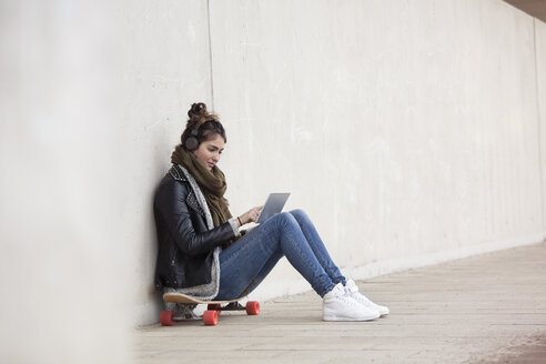 Junge Frau sitzt auf ihrem Longboard mit digitalem Tablet und hört Musik mit Kopfhörern - RBF002889