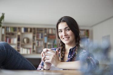 Porträt einer lächelnden jungen Frau mit Kaffeetasse, die sich zu Hause entspannt - RBF002894