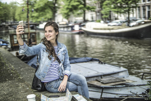Niederlande, Amsterdam, weibliche Touristin, die sich vor einer Gracht ausruht und ein Selfie macht - RIBF000111