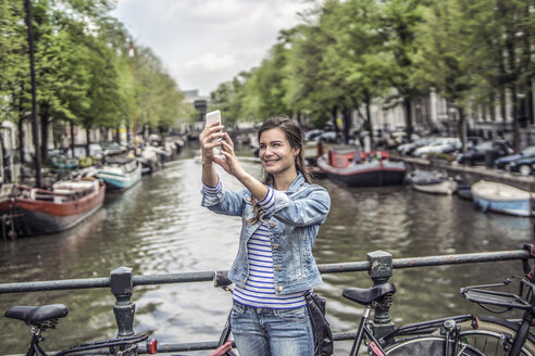 Niederlande, Amsterdam, weibliche Touristin macht ein Selfie mit Smartphone vor dem Stadtkanal - RIBF000080