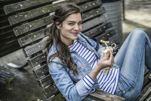 Niederlande, Amsterdam, lächelnde Frau sitzt auf einer Bank und isst Pommes frites - RIBF000112