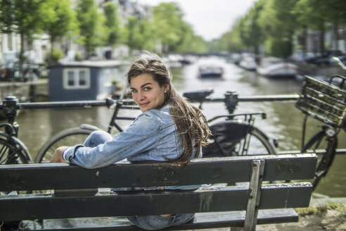 Niederlande, Amsterdam, lächelnde Frau sitzt auf einer Bank vor einer Gracht - RIBF000099