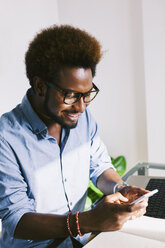 Smiling young man at home office looking at smartphone - EBSF000645