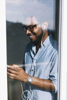Lächelnder junger Mann hinter einer Fensterscheibe, der Musik von seinem Smartphone hört - EBSF000652