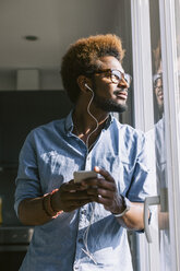 Junger Mann, der mit seinem Smartphone Musik hört und aus dem Fenster schaut - EBSF000655