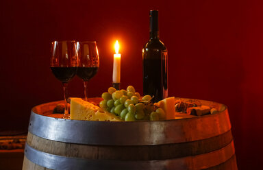 Zwei Gläser Rotwein, Flasche, Weintrauben, Käse und brennende Kerze auf einem Weinfass in einem Weinkeller - JTF000665