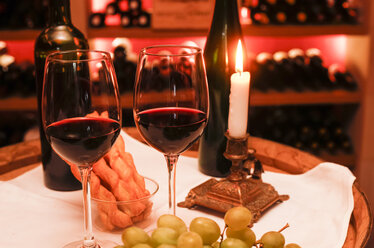 Zwei Gläser Rotwein, Weintrauben, Käsestangen und eine brennende Kerze in einem Weinkeller - JTF000666
