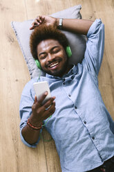 Junger afroamerikanischer Mann mit Kopfhörern und Smartphone auf dem Boden liegend - EBSF000612