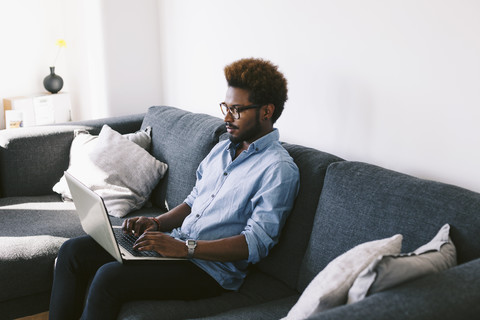 Junger afroamerikanischer Mann sitzt auf einer Couch und benutzt einen Laptop, lizenzfreies Stockfoto