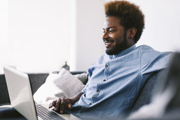 Junger afroamerikanischer Mann sitzt auf einer Couch und benutzt einen Laptop - EBSF000620