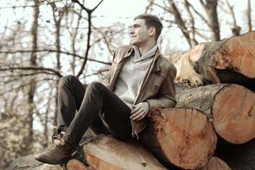Young man sitting on stack of tree trunks - GDF000734
