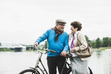 Happy senior couple standing at water's edge with bicycle - UUF004498