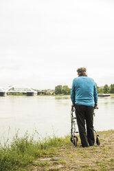 Rückenansicht eines älteren Mannes, der mit einer Gehhilfe auf Rädern am Wasser steht - UUF004506