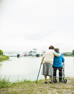 Rückenansicht eines älteren Ehepaars, das mit Gehstock und Rollator am Wasser steht - UUF004512