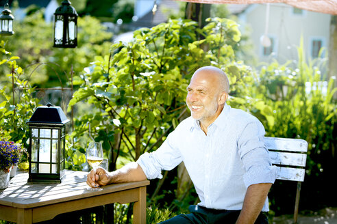 Portrait of happy man sitting with glass of white wine relaxing in the garden - MAEF010641