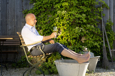 Mann mit einem Glas Weißwein nimmt ein Fußbad in einem Garten - MAEF010644