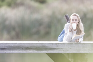 Frau mit Buch trinkt Kaffee auf einem Steg am See - ZEF005779