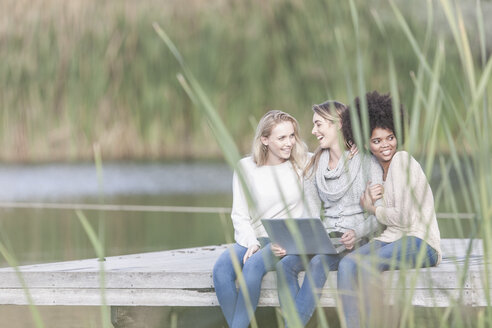 Three women with laptop relaxing on a jetty at lake - ZEF005780