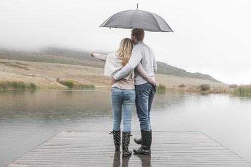 Back view of couple standing arm in arm in the rain on wooden boardwalk with umbrella - ZEF006220