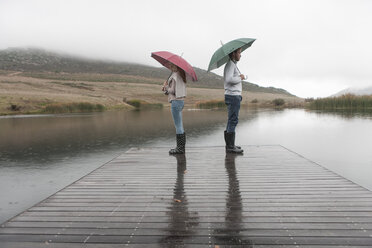 Couple standing in the rain on wooden boardwalk with umbrellas - ZEF006222