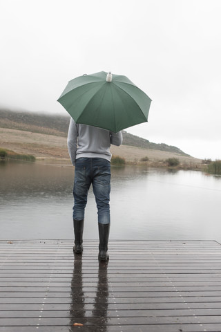 Mann steht im Regen auf einer Holzpromenade mit grünem Regenschirm, lizenzfreies Stockfoto