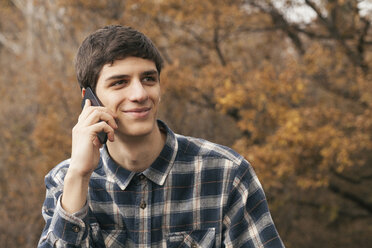 Porträt eines lächelnden jungen Mannes, der mit seinem Smartphone in einem Park telefoniert - BZF000163