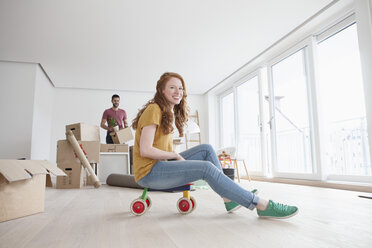 Junges Paar zieht in eine neue Wohnung ein, Frau sitzt auf einem Spielzeugwagen - RBF002851