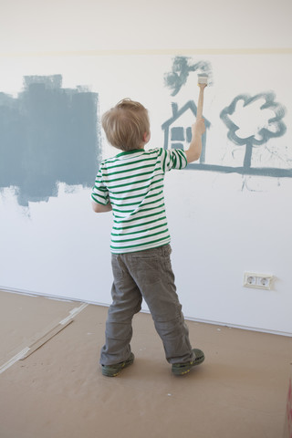 Kleiner Junge malt Haus und Baum an die Wand seines neuen Zimmers, lizenzfreies Stockfoto