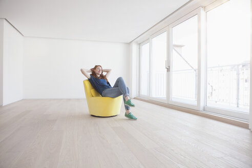 Young woman sitting on yellow armchair in her empty living room - RBF002752