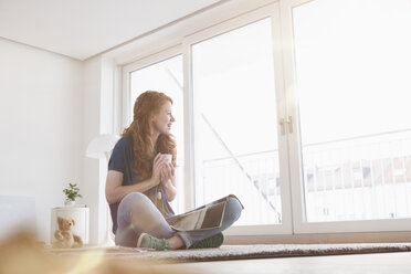 Young woman sitting on the floor of her living room looking through window - RBF002829