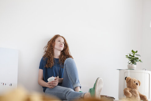 Tagträumende junge Frau sitzt auf dem Boden ihres Wohnzimmers - RBF002831