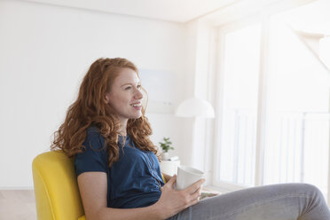 Glückliche junge Frau mit einer Tasse Kaffee in ihrem Wohnzimmer sitzend - RBF002754